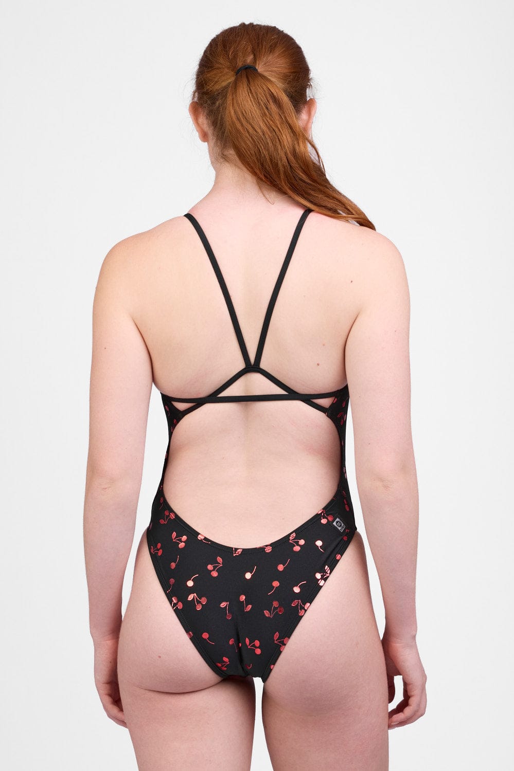 Female model wearing the Women’s Perry Swim Onesie in the Cherry Burst print, standing in front of a grey background, shown from back view. 